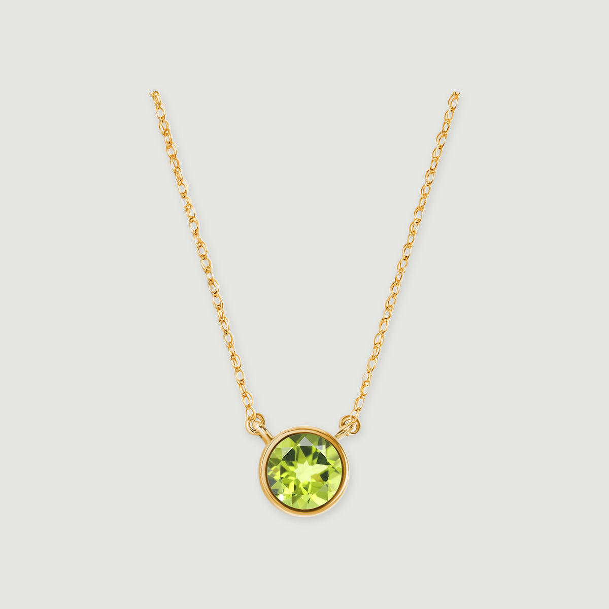 A round green peridot in a yellow gold bezel hangs between two fine chain sections on a light gray background.
