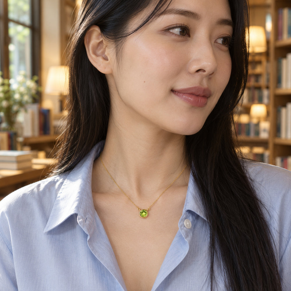 A woman with long black hair wears a yellow gold peridot necklace above a pale blue shirt in a softly lit bookshop.