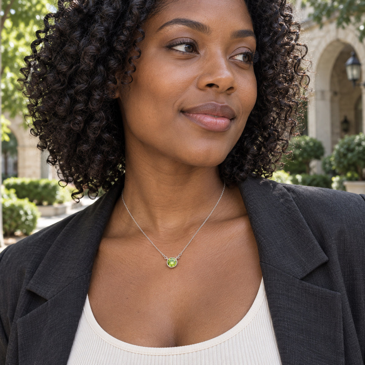 A woman with dark curls wears a white gold peridot necklace above a cream top and charcoal jacket in a leafy courtyard.