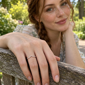 A rose gold diamond ring is shown on a hand resting on a weathered wooden bench in a garden.