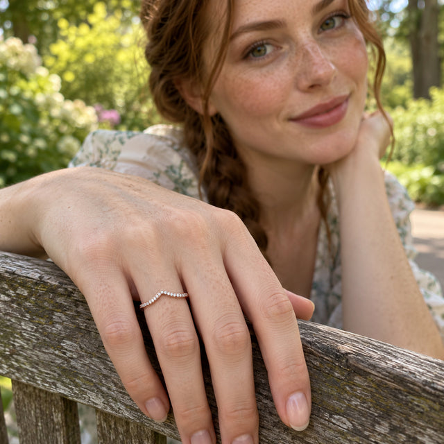 A rose gold diamond ring is shown on a hand resting on a weathered wooden bench in a garden.