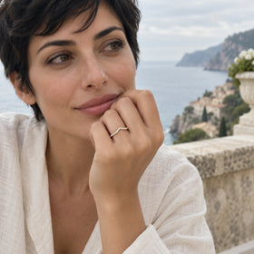 A woman wears a yellow gold diamond ring on her raised hand with a coastal terrace behind her.