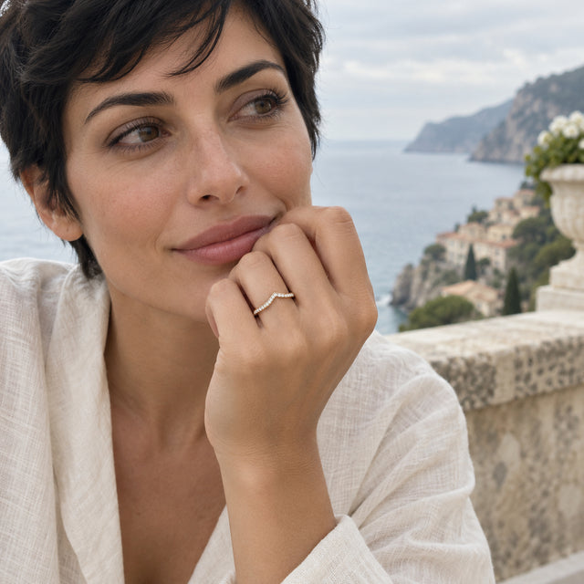 A woman wears a yellow gold diamond ring on her raised hand with a coastal terrace behind her.