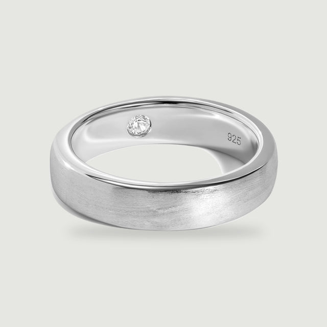 A brushed silver ring with a polished rim and a small clear stone inside rests on a light gray surface.