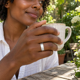 A woman wears the brushed silver band while holding a ceramic cup at a leafy outdoor cafe.