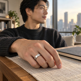 A man rests his hand with the silver band on a linen-covered wooden desk beside a window.