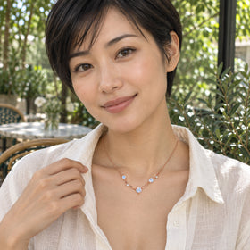 A woman with short dark hair wears a rose gold moonstone necklace over an open white shirt at a leafy cafe.