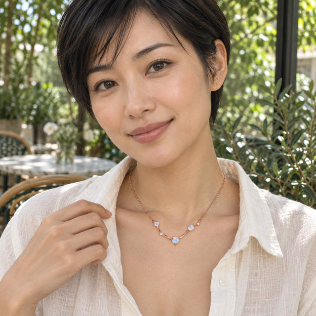 A woman with short dark hair wears a rose gold moonstone necklace over an open white shirt at a leafy cafe.
