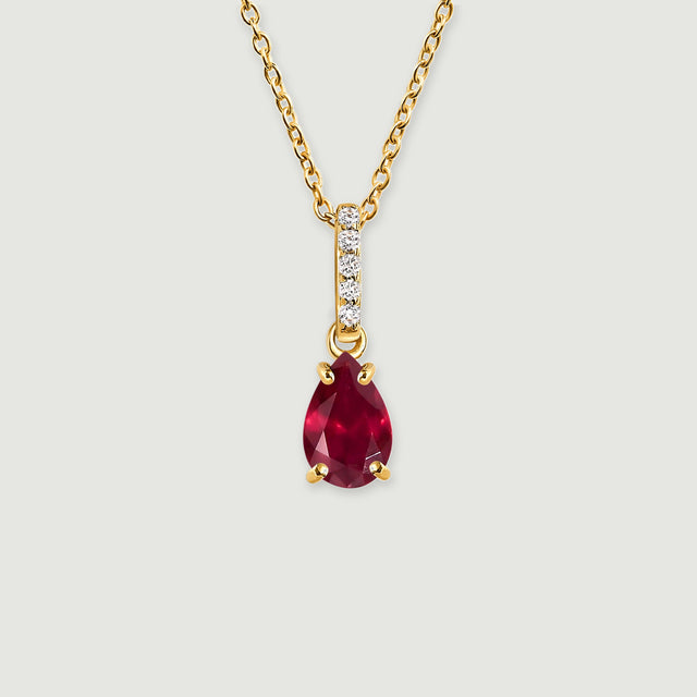 A yellow gold necklace has a pear-shaped red gemstone suspended beneath a vertical row of five clear stones on a light gray background.