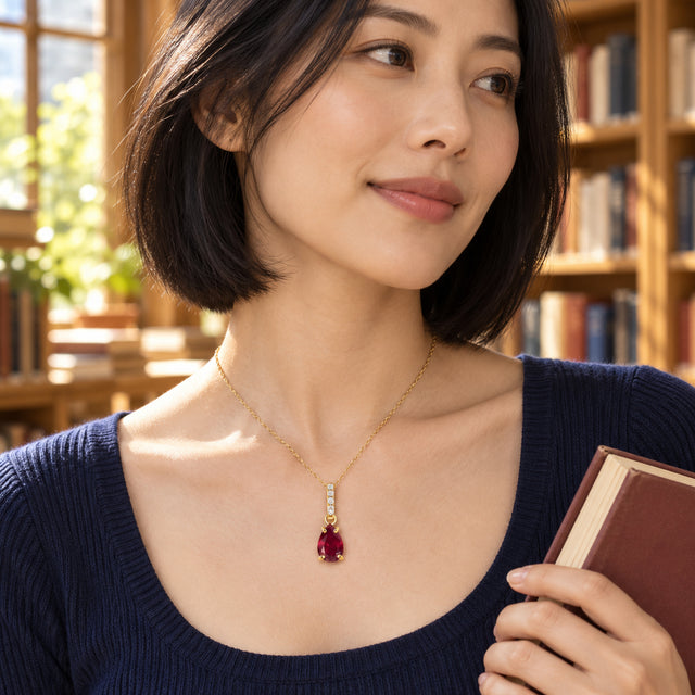 A woman in a navy knit top wears a gold red-gemstone necklace while holding a book beside wooden shelves.