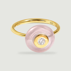 A yellow gold ring with a translucent pink circular stone and a clear center gem rests on a pale gray surface.