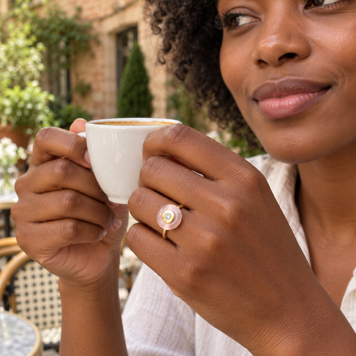A yellow gold ring with a round pink stone is worn on a hand holding a white coffee cup in a leafy courtyard.