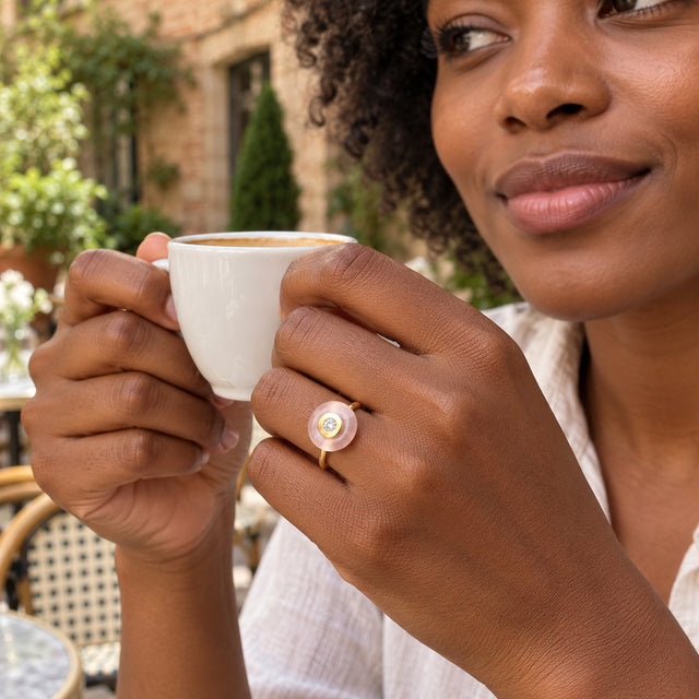 A yellow gold ring with a round pink stone is worn on a hand holding a white coffee cup in a leafy courtyard.
