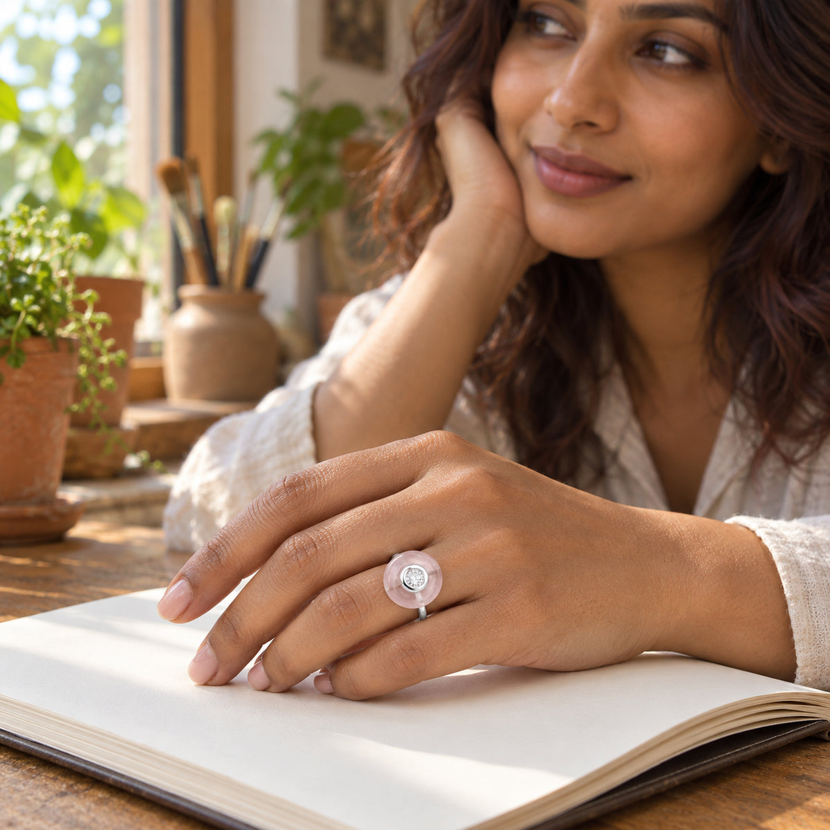 A silver ring with a pink circular stone and a clear center gem is shown on a hand resting on an open blank sketchbook.