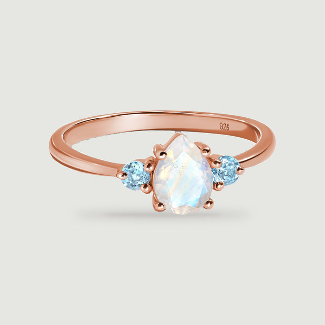 A rose gold ring has a pear-shaped pale moonstone between two round light blue stones.