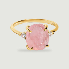 A yellow gold ring with a rough pink stone and two round clear side stones rests on a light gray surface.