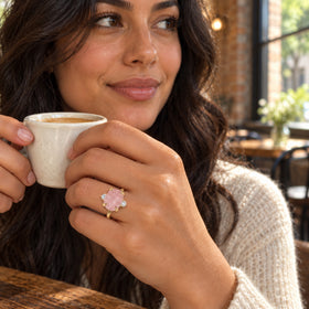 A yellow gold ring with a rough pink stone is worn on a hand holding a ceramic cup in a cafe.