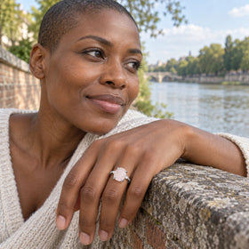 A white gold ring with a pale pink stone is shown on a hand resting on a riverside stone wall.