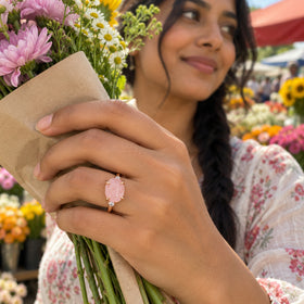 A rose gold ring with a pink stone is worn on a hand holding a paper-wrapped bouquet at a flower market.