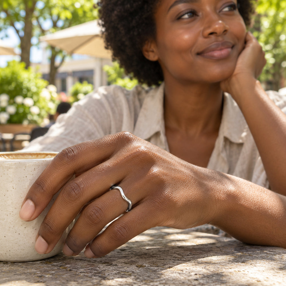A silver contoured ring is worn on a hand beside a ceramic cup at an outdoor cafe.