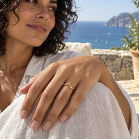 A yellow gold band is worn on a hand resting on white linen with the sea in the background.