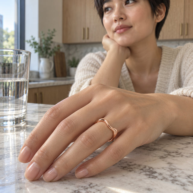 A rose gold contoured ring is worn on a hand resting on a marble kitchen counter beside a glass of water.