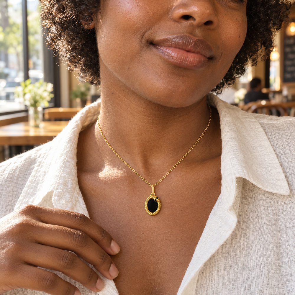 A woman in an ivory linen shirt wears the yellow gold black-stone pendant in a sunlit cafe.