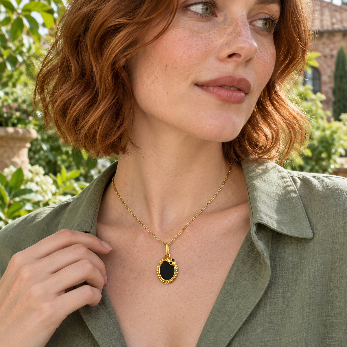 A woman in a sage green blouse wears the yellow gold oval pendant beside leafy plants.