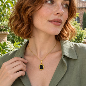 A woman in a sage green blouse wears the yellow gold oval pendant beside leafy plants.