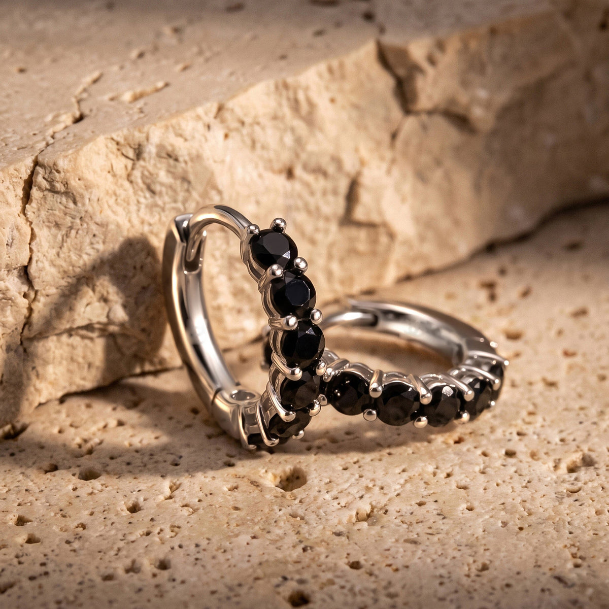 Two silver-colored black-stone hoops lean together on a textured beige stone surface.