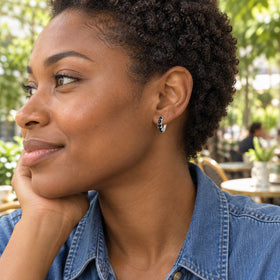 A woman with short curly hair wears a silver-colored black-stone hoop at a leafy outdoor cafe.