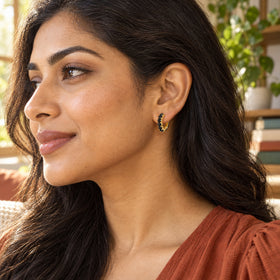 A woman with long dark hair wears a gold-colored black-stone hoop beside a sunlit window.