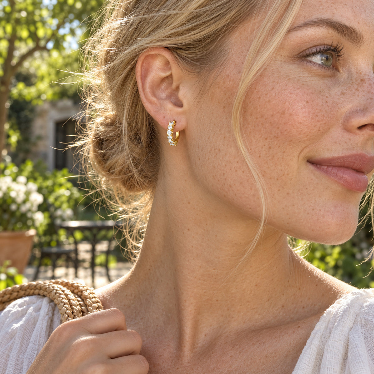 A woman with blonde hair wears a yellow gold-tone white topaz hoop in a sunlit garden.