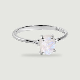 A sterling silver ring holds a rough pale moonstone between two round side stones on a light gray background.