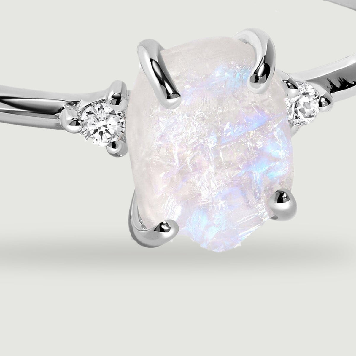 A close view shows blue flashes across the rough moonstone, four silver prongs and the clear stones beside it.