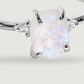 A close view shows blue flashes across the rough moonstone, four silver prongs and the clear stones beside it.