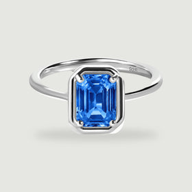 A polished silver ring with a rectangular blue gemstone stands on a light gray background.
