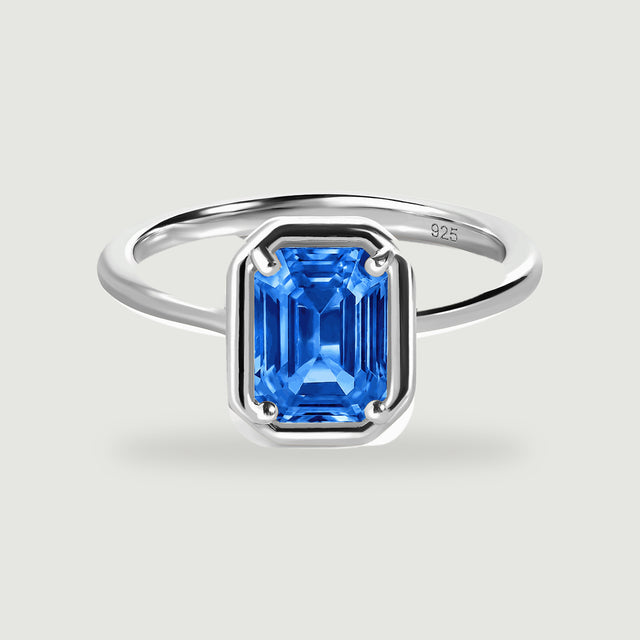 A polished silver ring with a rectangular blue gemstone stands on a light gray background.