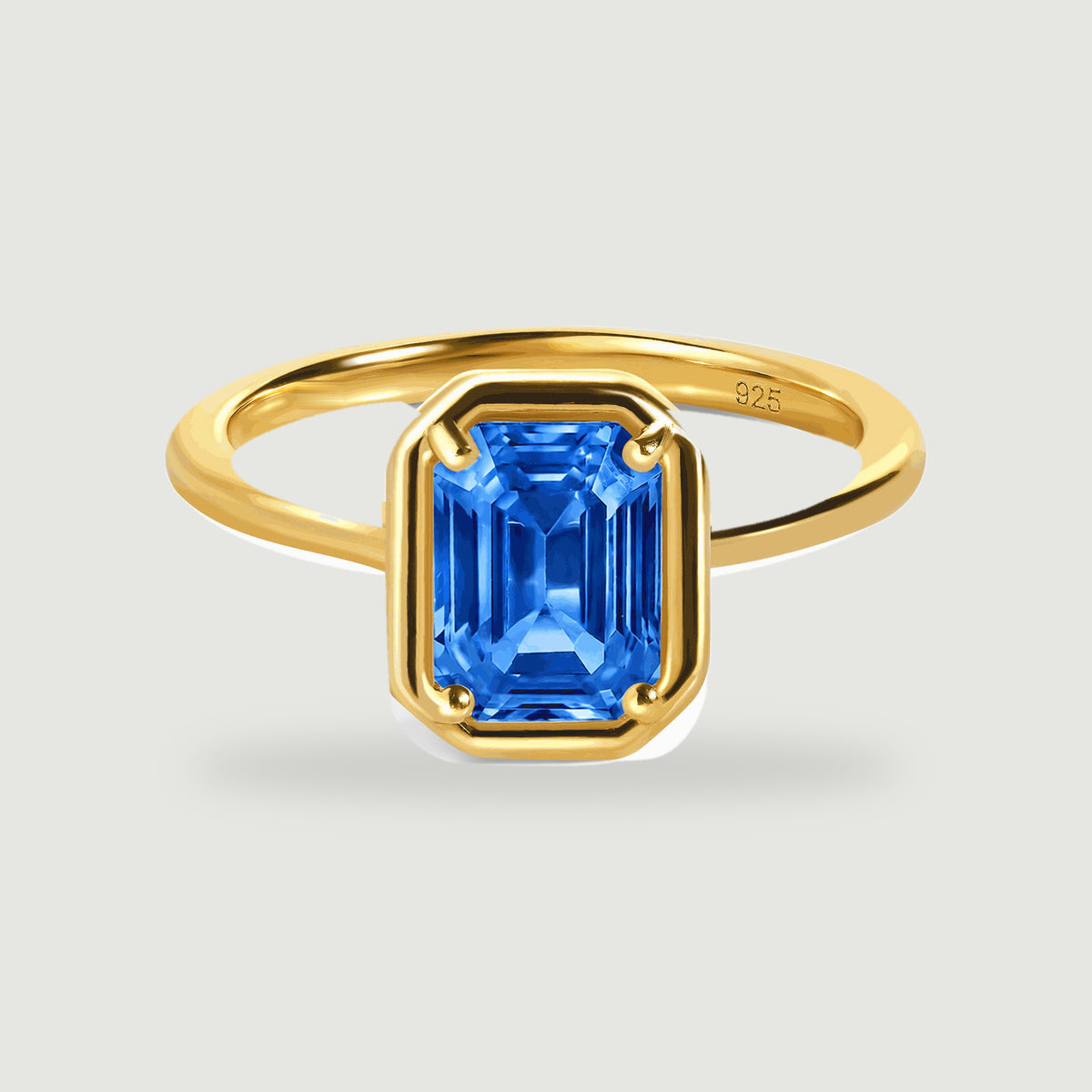 A yellow gold ring holds a blue emerald-cut gemstone above a soft contact shadow.