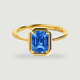 A yellow gold ring holds a blue emerald-cut gemstone above a soft contact shadow.