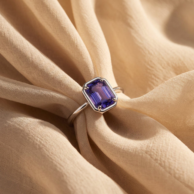 A silver ring with a violet faceted stone rests between folds of light linen.