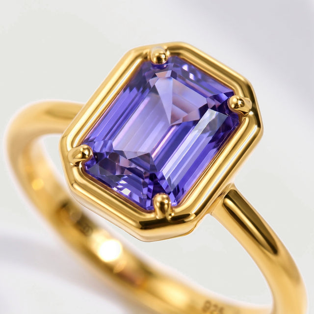 A violet emerald-cut gemstone is shown close up in a yellow gold four-prong setting.