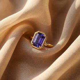 A yellow gold ring with a violet rectangular stone lies on softly folded linen.
