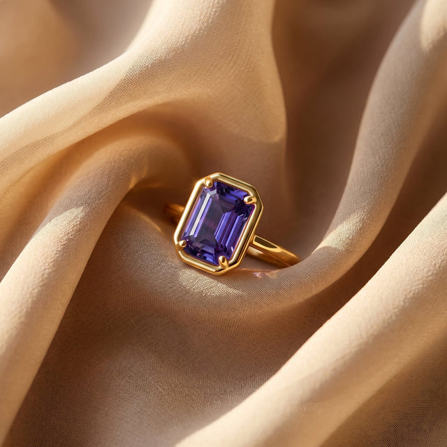 A yellow gold ring with a violet rectangular stone lies on softly folded linen.