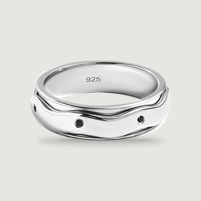 A polished silver ring with small round black stones and a wavy outer band sits on a light gray surface.