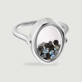 A polished sterling silver ring holds black and blue gray gems inside a clear oval chamber on a light gray surface.