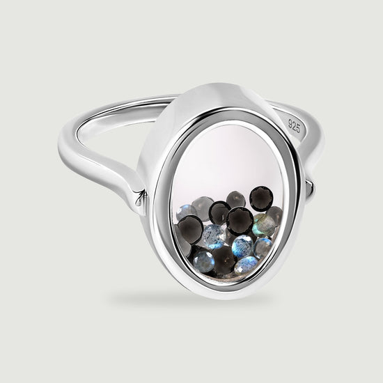 A polished sterling silver ring holds black and blue gray gems inside a clear oval chamber on a light gray surface.