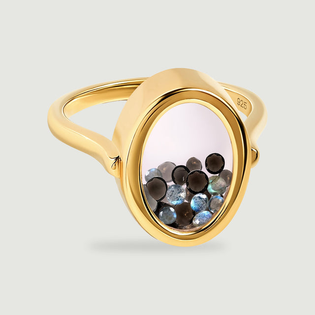 A yellow gold vermeil ring with a clear oval window contains small dark and iridescent gems against a pale gray background.