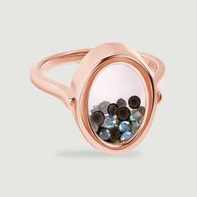 A rose gold vermeil ring displays loose black and blue gray stones beneath its clear oval cover on a soft gray surface.