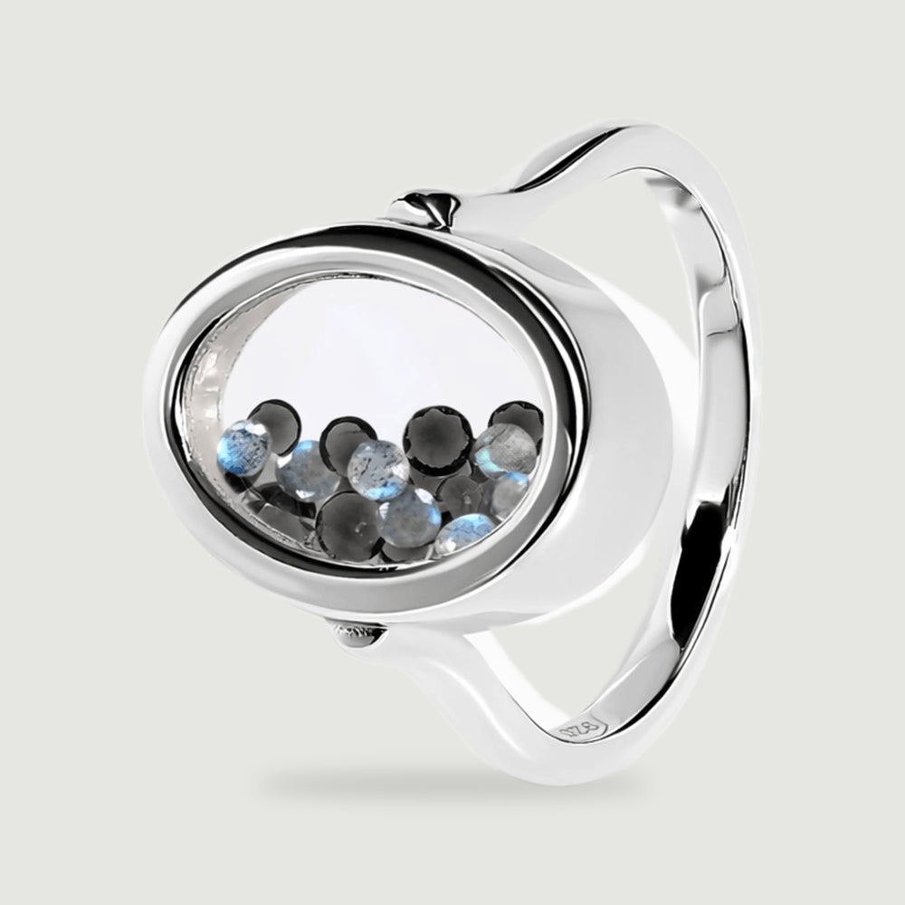 An angled sterling silver ring reveals its oval chamber, reflective band, and small round black and blue gray gems.